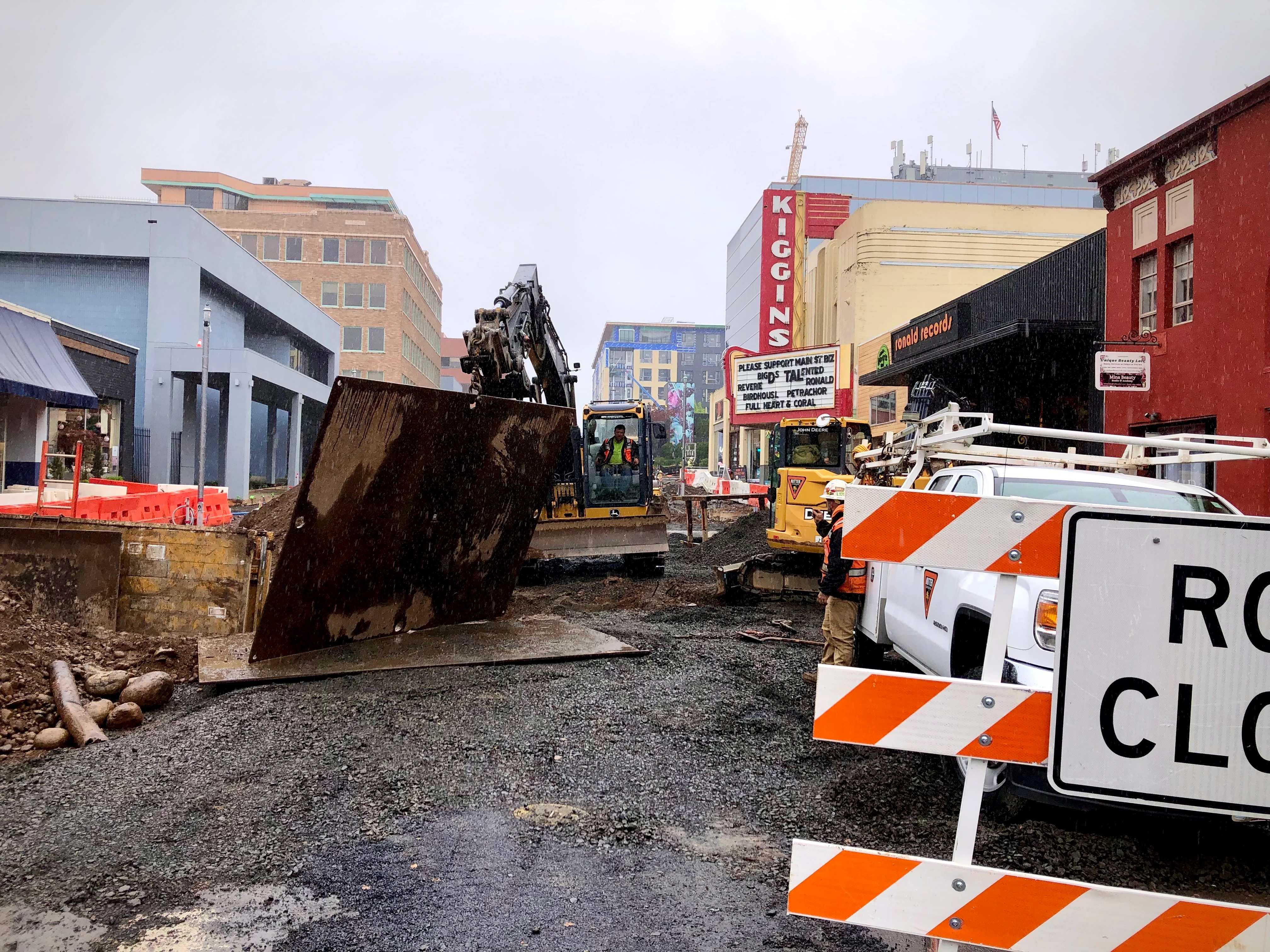 Main Street Vancouver under construction in the rain, April 2026, excavators working outside Ronald Records with the Kiggins Theatre marquee reading Please Support Main St Biz